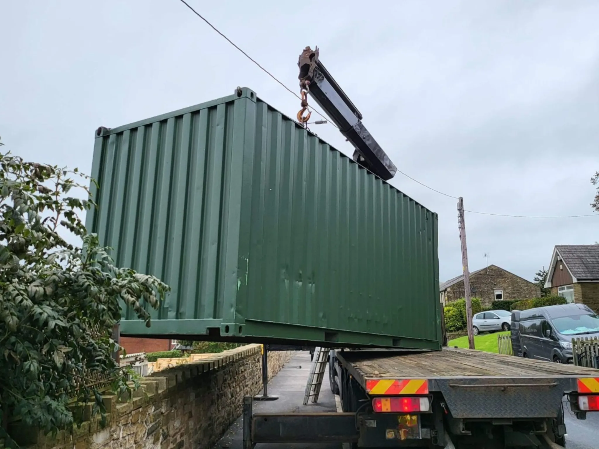 20ft new one trip shipping container delivered by HIAB crane over a hedge on a residential street