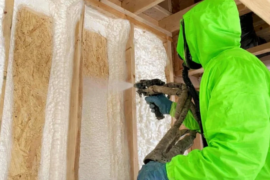 Worker applying spray foam insulation between shipping container timber studs while wearing a bright green protective suit and blue gloves inside a timber-framed structure.
